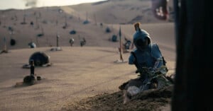 A desert landscape with scattered helmets and armor pieces on sand mounds, resembling makeshift graves. In the foreground, a blue helmet and chest plate are placed among stones and small branches.