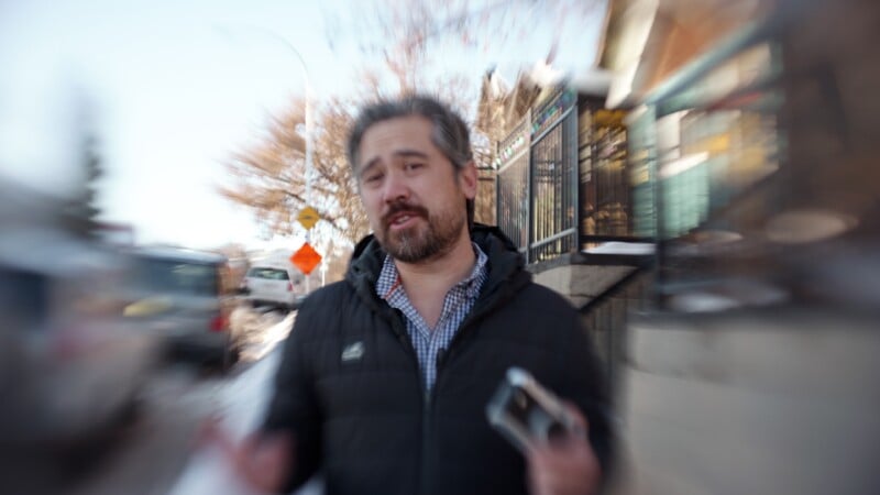 A man with gray hair and a beard stands outdoors in winter, wearing a dark jacket and holding a phone. The background, including trees, cars, and buildings, is blurred with a zoom effect.