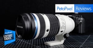 A large OM System camera lens attached to a camera body is displayed on a grid-patterned surface. Overlay text reads "PetaPixel Reviews" and "PetaPixel Editors' Choice.