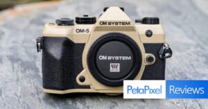 A beige OM SYSTEM OM-5 camera body with a black grip is displayed on a marble surface. The lens mount cap shows “OM SYSTEM Micro.” The Petapixel Reviews logo appears in the lower right corner.