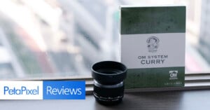 A camera lens and a box labeled "OM System Curry" are placed on a windowsill. The background shows a blurred cityscape, while the foreground features a "PetaPixel Reviews" banner.