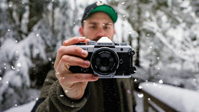 A person holding a silver and black camera toward the viewer, standing outdoors in a snowy forest. Snowflakes are falling, and the background is blurred with snow-covered trees.