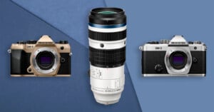 Two OM System OM-5 cameras, one gold and one silver, are displayed on either side of a large white telephoto lens labeled 150-600mm, all set against a blue geometric background.