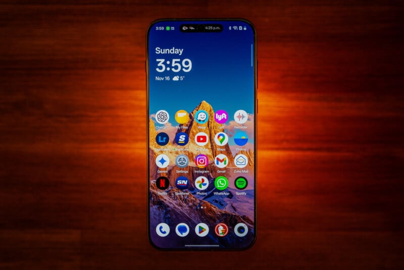 A smartphone with a colorful mountain wallpaper is displayed on a wooden surface. Its screen shows various app icons, the time (3:59), date (Sunday, Nov 16), and weather.
