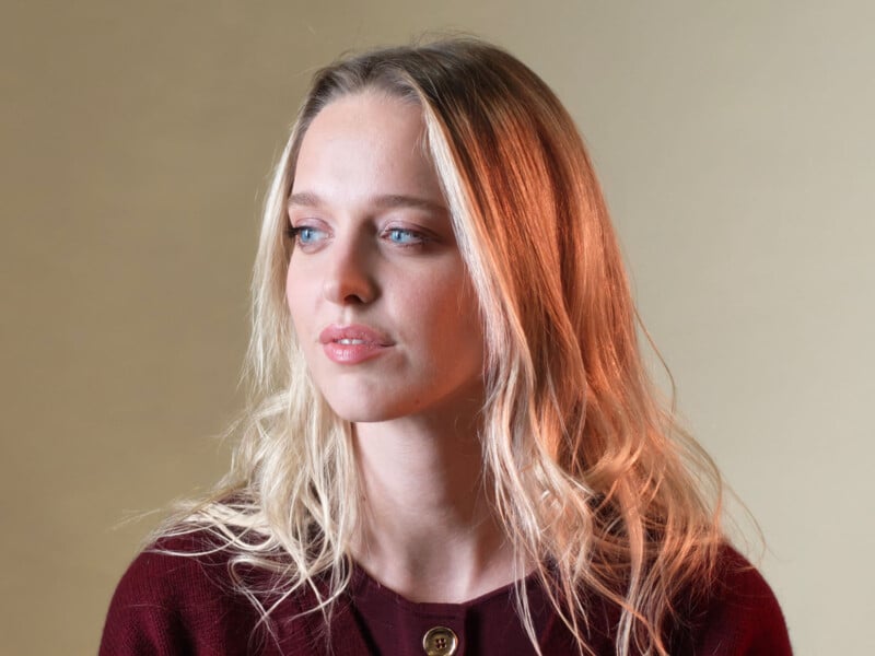 A young woman with long blonde hair and blue eyes looks off to the side. She is wearing a maroon sweater and is posed against a plain beige background with soft lighting.
