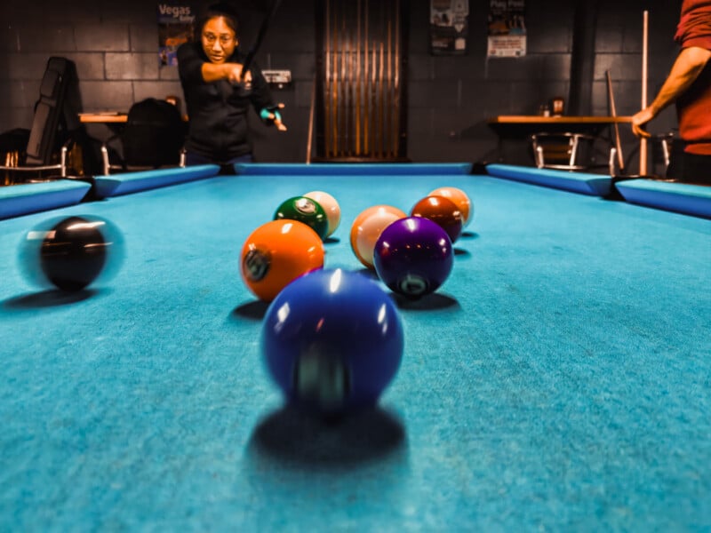 A person is breaking in a game of pool, striking the cue ball towards a racked set of colorful billiard balls on a bright blue pool table. Another person stands to the side. The scene is indoors, with a dark background.