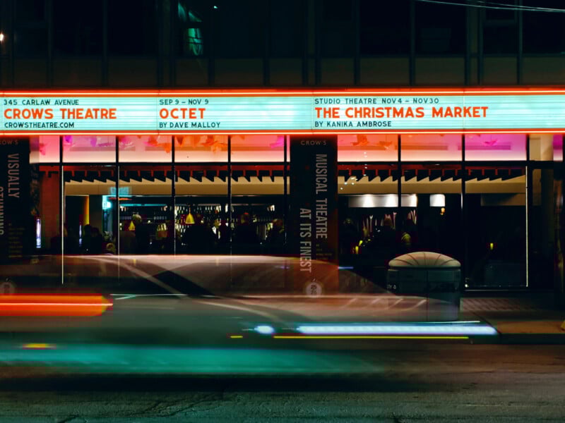 A theater at night with a bright marquee advertising shows including "Octet" and "The Christmas Market." Light trails from passing cars are visible in front of the building.