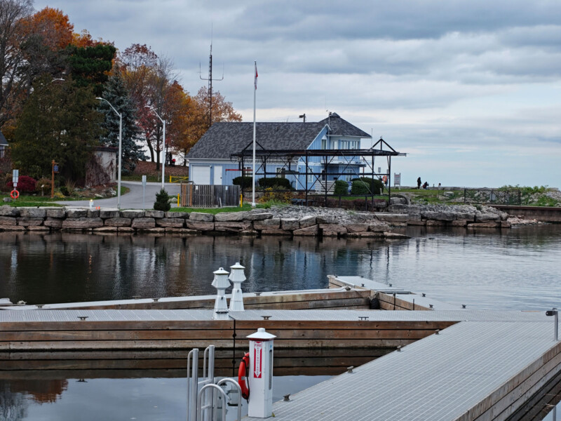 A small marina with empty docks, calm water, and a blue boathouse on the shore. Autumn trees and cloudy skies are in the background. A stone wall separates the marina from a road along the shore.