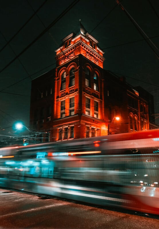 A historic red-brick building is lit up at night, with a brightly illuminated streetcar passing by in the foreground, creating light streaks and motion blur on a city street.