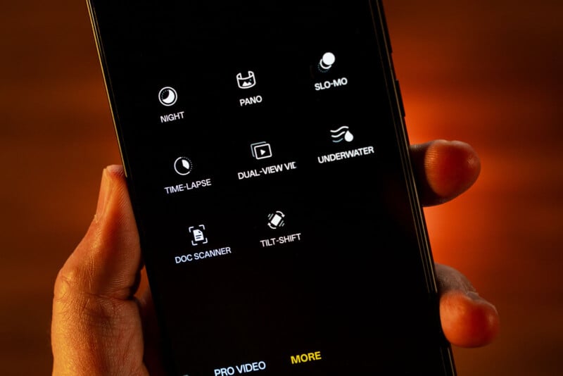 A hand holds a smartphone showing a camera app menu with icons for various modes, including Night, Pano, Slo-mo, Time-lapse, Dual-view, Underwater, Doc Scanner, and Tilt-shift.