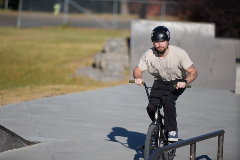 A man wearing a helmet and light-colored shirt rides a BMX bike at a skatepark, approaching a metal rail on a sunny day.