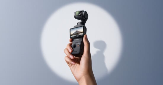 A hand holds up a compact handheld camera with a small screen displaying a landscape image. The camera has a gimbal-mounted lens and is set against a plain, softly lit background.