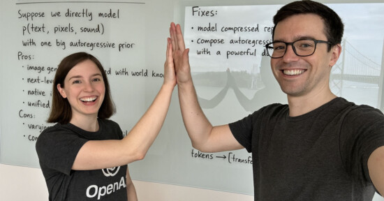Two people smiling as they high-five in front of a whiteboard with handwritten notes. The whiteboard includes points under "Pros" and "Fixes." Both are wearing casual dark t-shirts.