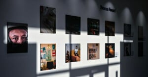 A sunlit gallery wall displays a grid of photographs, with shadows from window panes creating patterns. The photos include a close-up of a person's eyes, various scenes, and interior shots. A sign reads "Bronze Award.