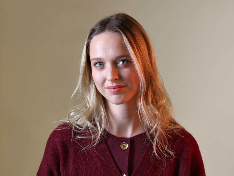 A young woman with light skin and long blonde hair wears a maroon sweater and smiles softly at the camera against a plain beige background.