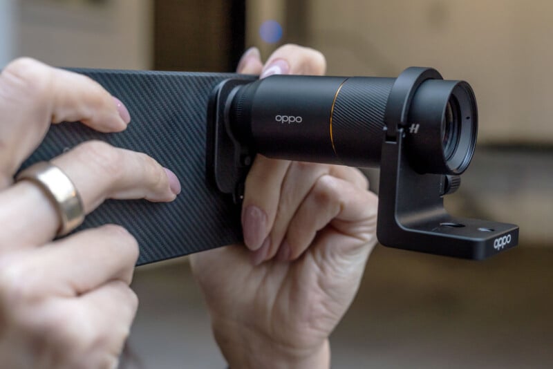 A person holds a smartphone with a textured black case, using an attached Oppo telephoto lens for enhanced photography. The person’s hands have polished nails and wear rings. The background is blurred.
