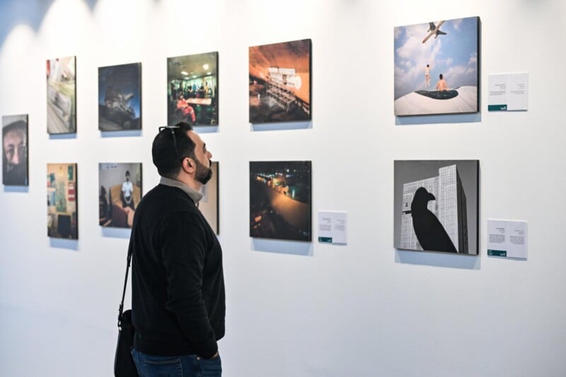 A man with a beard stands in front of a white wall, looking at a display of square photographs in an art gallery. The images include various scenes and are accompanied by small information cards.