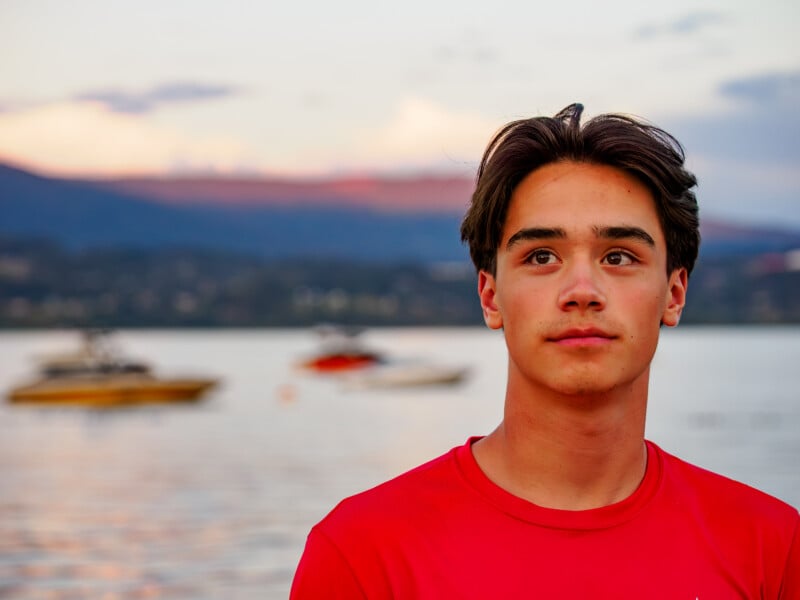 A teenage boy with dark hair wearing a red shirt stands by a calm lake at sunset, with blurred boats and mountains in the background.