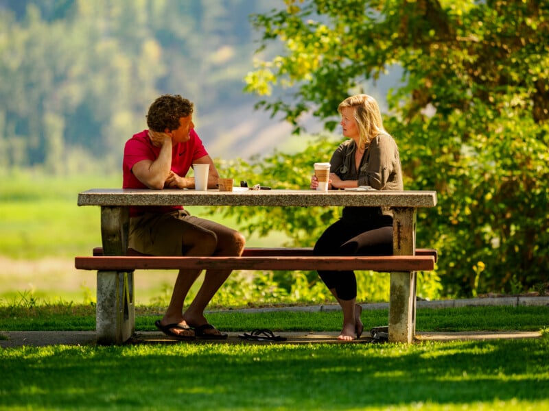 A man and a woman sit across from each other at a stone picnic table in a park, having a conversation and holding coffee cups, surrounded by green grass and trees in sunlight.