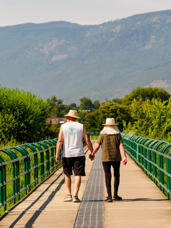 An older couple wearing hats walks hand-in-hand down a sunny, green-lined bridge with mountains visible in the background.
