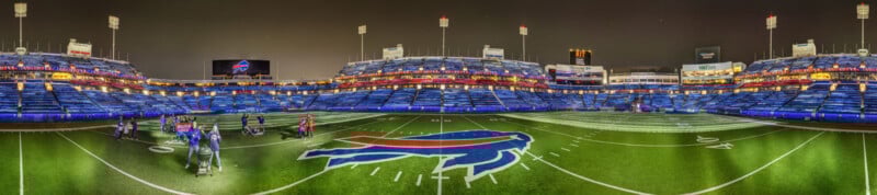 A panoramic view of a brightly lit football stadium at night, featuring the Buffalo Bills logo at the center of the field and several people gathered on the turf. The stands are mostly empty.