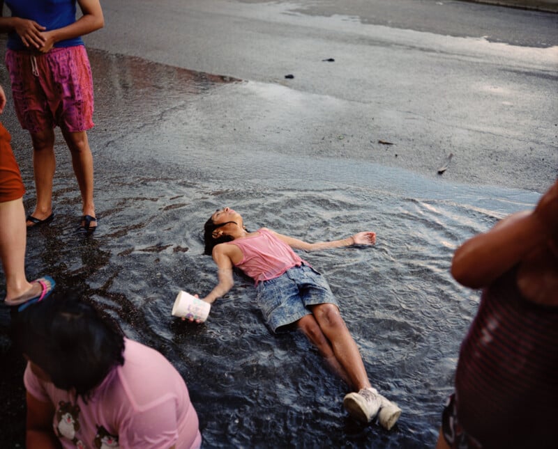 A child in shorts and a pink shirt lies on their back in a puddle on a wet street, holding a white cup, while other people stand nearby, mostly out of frame.