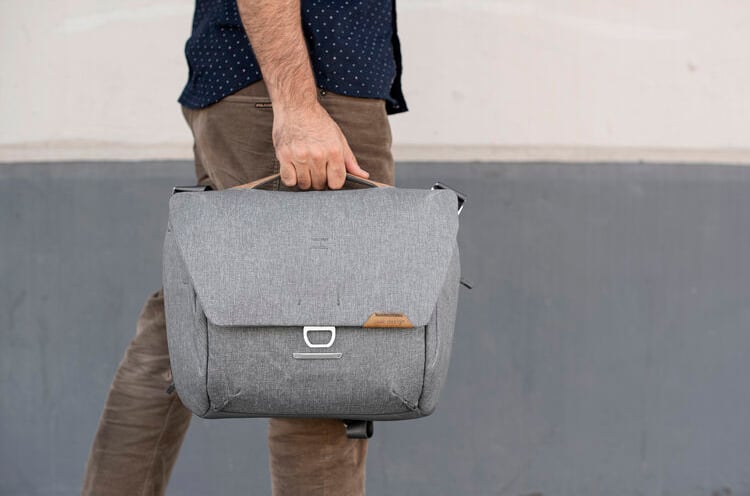 A person wearing brown pants and a navy shirt holds a gray messenger bag by its top handle, standing in front of a two-tone gray and white wall.