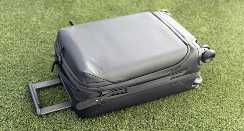 A black hard-shell suitcase with an extended handle lies on green grass. The suitcase has wheels and zippers, and is positioned horizontally.