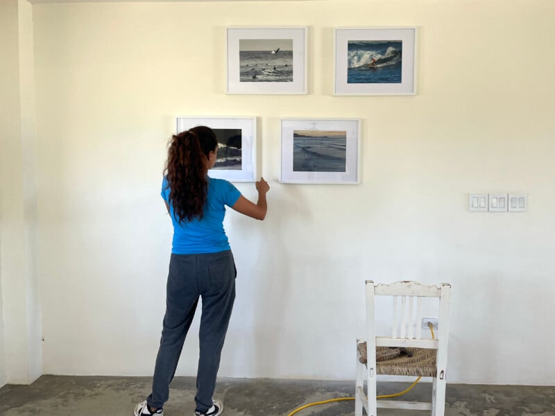 A woman with long brown hair, wearing a blue shirt and gray pants, hangs a framed photo on a white wall with three other framed pictures. A white chair and a yellow extension cord are nearby.