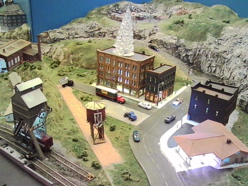 A detailed model train layout features buildings, roads, cars, a water tower, and rocky hills. A decorated tree sits atop a brick building, with tracks and a train car visible to the left.