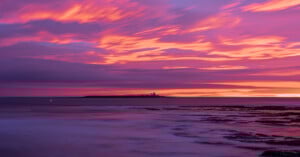 Vibrant sunset over a calm sea, with dramatic purple, pink, and orange clouds; a distant island with a lighthouse is silhouetted against the glowing sky, and gentle waves wash over rocky shoreline in the foreground.