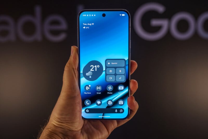 A hand holds a smartphone displaying a modern blue-themed home screen with weather, app icons, and widgets, including a "Gemini" widget. The blurred background shows partial text reading "Made by Google.