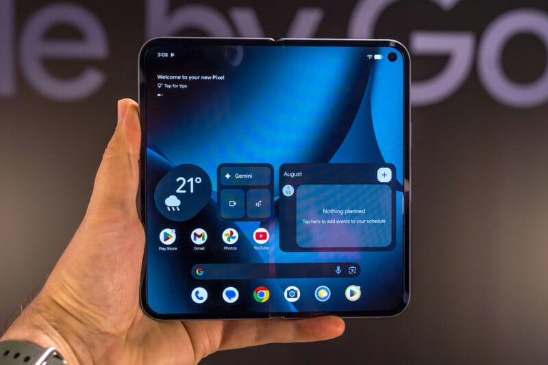 A hand holds an open foldable smartphone displaying a home screen with weather, calendar, and app widgets on a blue-themed background. The device has a large, seamless display with rounded corners.