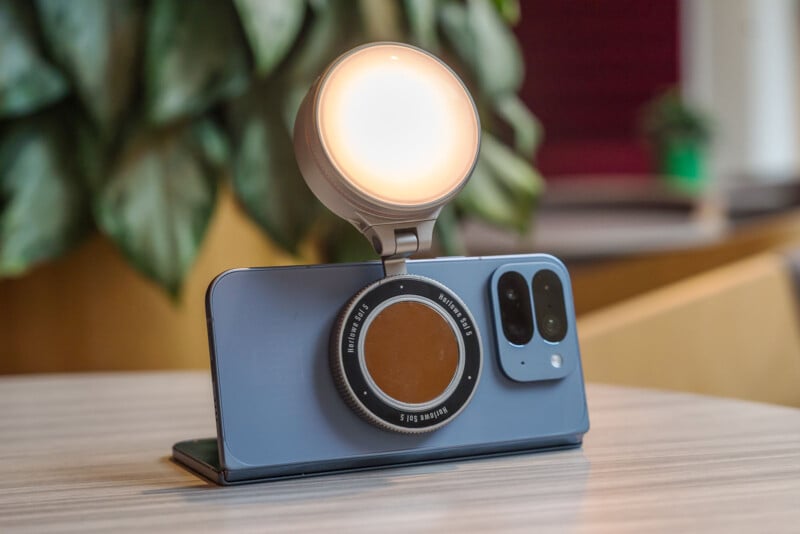 A smartphone in a blue case stands upright on a table with a circular, glowing light attachment mounted on the back. Blurred green leaves and indoor decor are visible in the background.