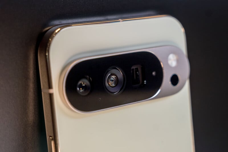 Close-up of the rear camera module on a modern smartphone with multiple lenses and sensors, set against a dark background. The phone has a glossy, light-colored finish.