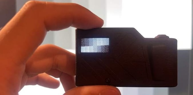 A hand holds a small black electronic device with a lit rectangular screen displaying white pixelated text and symbols against a blurred background.