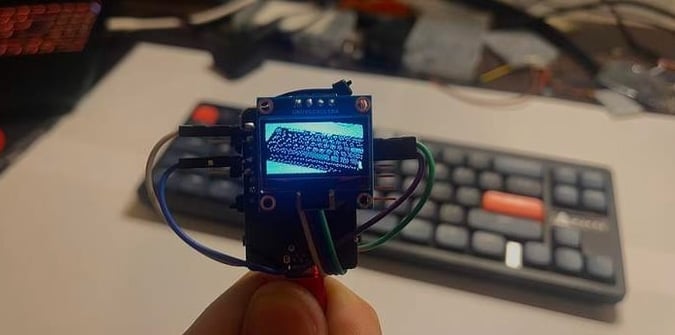 A hand holds a small electronic display showing a pixelated image of a bridge; a blurred keyboard and desk are visible in the background.