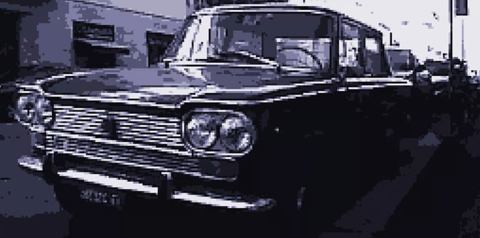 A vintage car with round headlights and a boxy design is parked on a city street, with other vehicles and buildings visible in the background. The image has a blue-tinted, low-resolution effect.