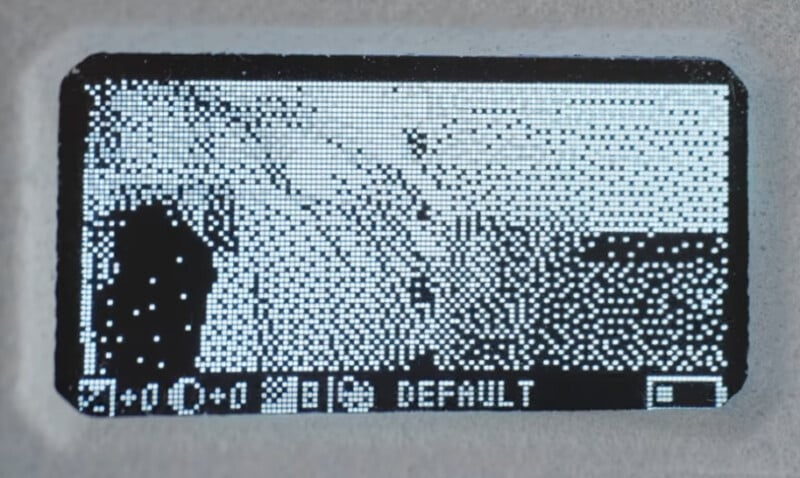 A pixelated grayscale display shows abstract shapes and patterns, with icons and the word "DEFAULT" at the bottom, resembling a digital device’s settings or status screen.