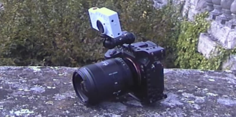 A black professional camera with a large lens sits on a stone surface outdoors, with a small white action camera mounted on top. Green foliage and stone railings are visible in the background.