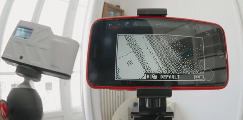 A smartphone mounted on a tripod displays a pixelated black-and-white camera view with the word "DEFAULT" on the screen, while a handheld camera is positioned nearby. Bright windows are visible in the background.