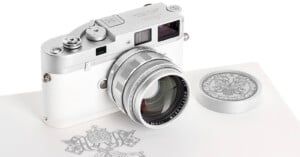 A silver and white Leica camera with a matching lens and cap, placed on a surface featuring an ornate emblem with detailed linework. The camera has elegant, classic design elements.