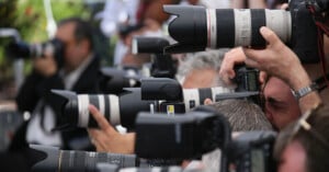 A group of photographers closely gathered, holding large professional cameras with telephoto lenses, aiming their cameras in the same direction, suggesting a press event or media gathering.