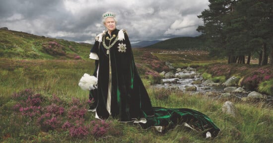 A person in regal attire with a crown stands in a scenic landscape, wearing a long, dark cape with white fur trim and holding a white fur accessory. The background features grassy fields, a small stream, and cloudy skies.