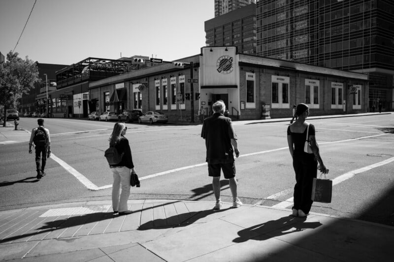 Four people stand at a street corner in a city, waiting to cross. Shadows are prominent on the sidewalk, and a large building with a mural is visible across the street. The scene is in black and white.