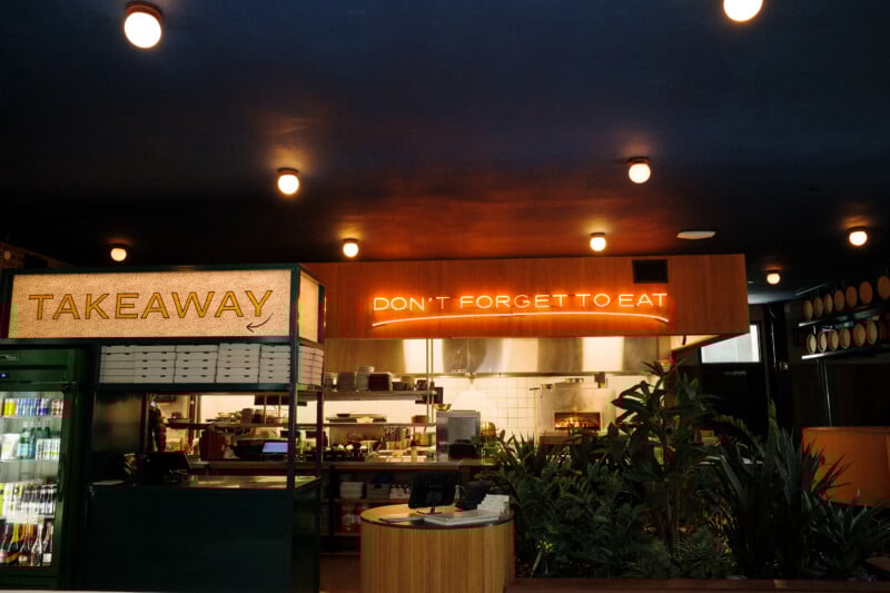 A restaurant counter with a TAKEAWAY sign on the left and a neon sign above the kitchen reading “DON’T FORGET TO EAT.” Indoor plants and shelves of drinks are visible in the brightly lit space.