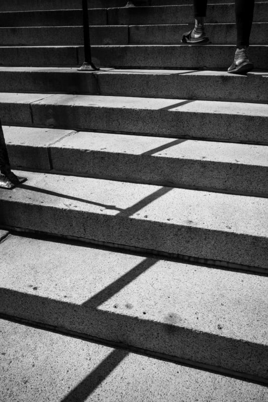 Black and white photo of concrete steps with shadows forming diagonal lines. Two people’s legs and feet are visible at the top, ascending the stairs. The lighting creates strong contrasts on the stair surfaces.
