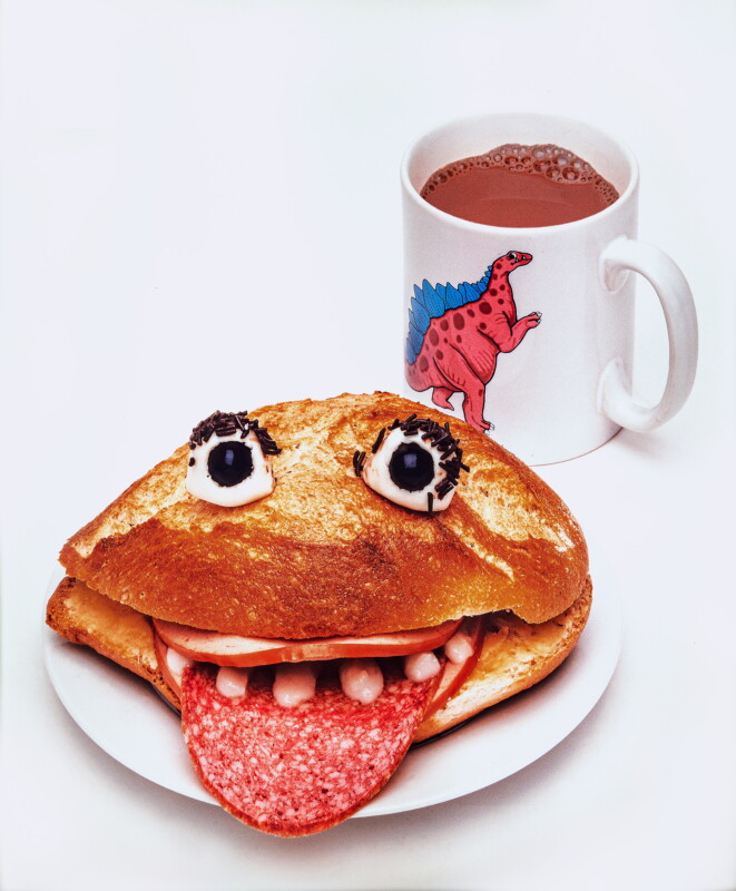 A sandwich decorated to look like a face with olive eyes, salami tongue, and cheese teeth sits on a plate next to a white mug with a cartoon dinosaur and hot chocolate inside.