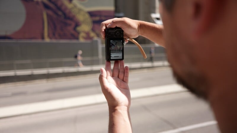 A person outdoors holds a camera with both hands, taking a photo of a mural across the street. The mural and a pedestrian are visible on the camera's screen. The scene is bright and sunlit.