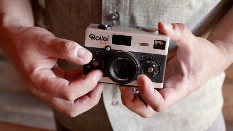 A person holding a vintage Rollei 35AF camera with both hands. The camera is silver and black with various dials and a lens in the center. The person is wearing a light-colored shirt in the background.